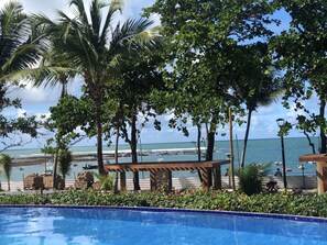 A pool area with a view of the ocean, surrounded by palm trees and a wooden pergola. - Apartamento Enseada Praia do Forte (Mata de São João)