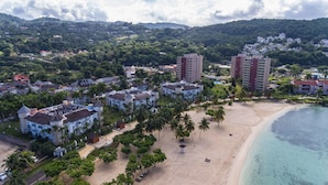 Vista aérea de un complejo turístico costero con villas frente a la playa, una playa de arena y colinas exuberantes al fondo.