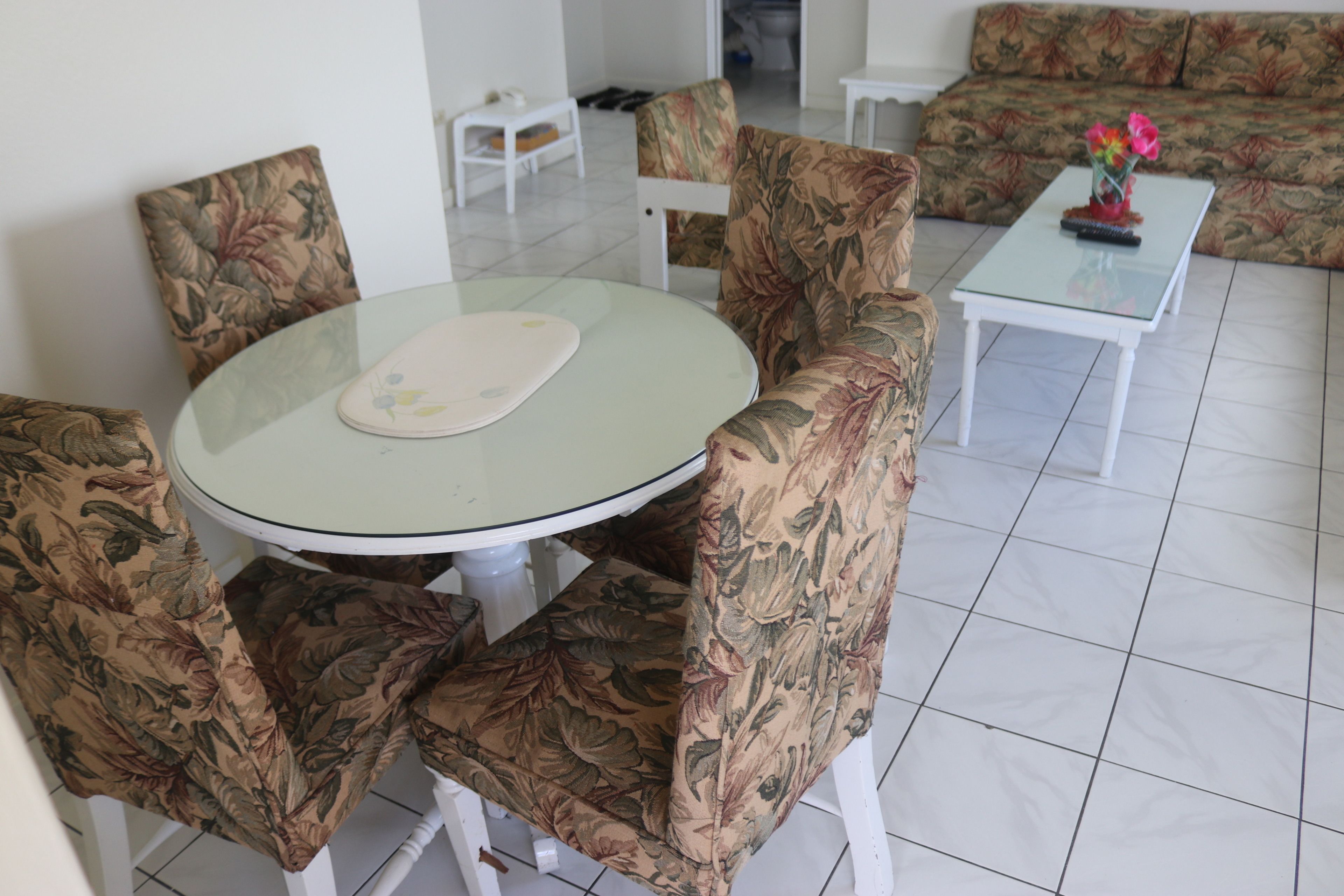 Uma área de refeições com uma mesa redonda de vidro, cadeiras com padrão floral, um sofá branco e uma pequena mesa com um vaso de flores.