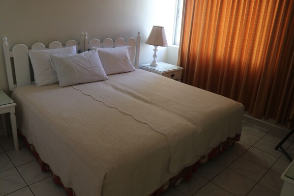 A neatly made bed with a white headboard, a bedside table with a lamp, and a window with curtains. - Angel Fish Beach Suites - Turtle Tower (Ocho Rios)