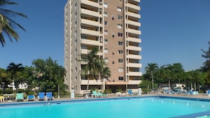 Una piscina con sillas de descanso y palmeras, rodeada por un edificio alto con varios balcones.