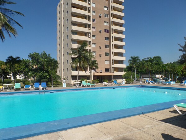 A swimming pool with lounge chairs and palm trees, surrounded by a tall building with multiple balconies.