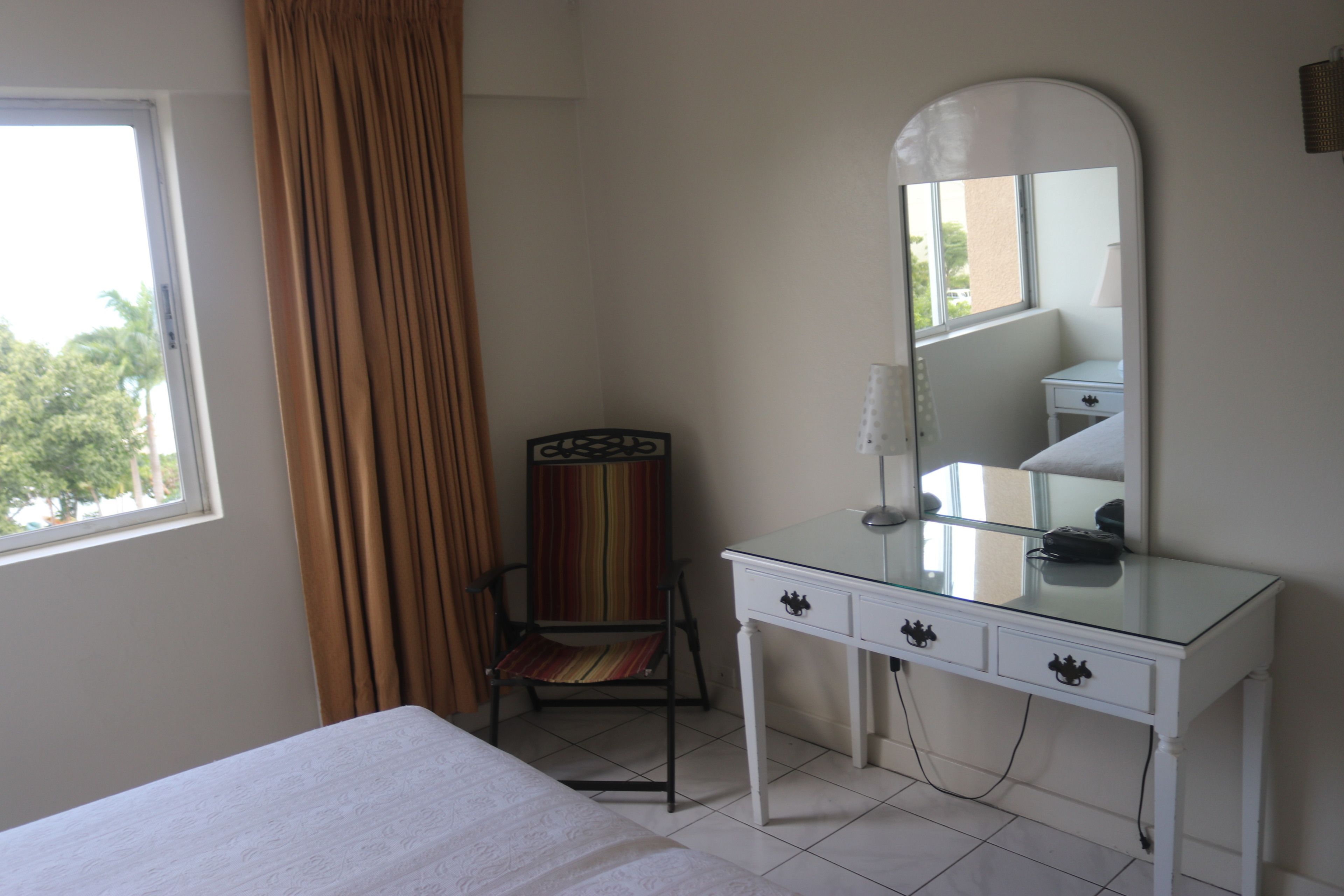 A hotel room with a bed, a white dresser with drawers, a chair, a large mirror, and a window with curtains.