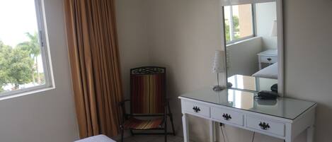 A hotel room with a bed, a white dresser with drawers, a chair, a large mirror, and a window with curtains.