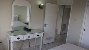 Un dormitorio con cama, una mesita de noche blanca con cajones, un espejo y una puerta que da a otra habitación.