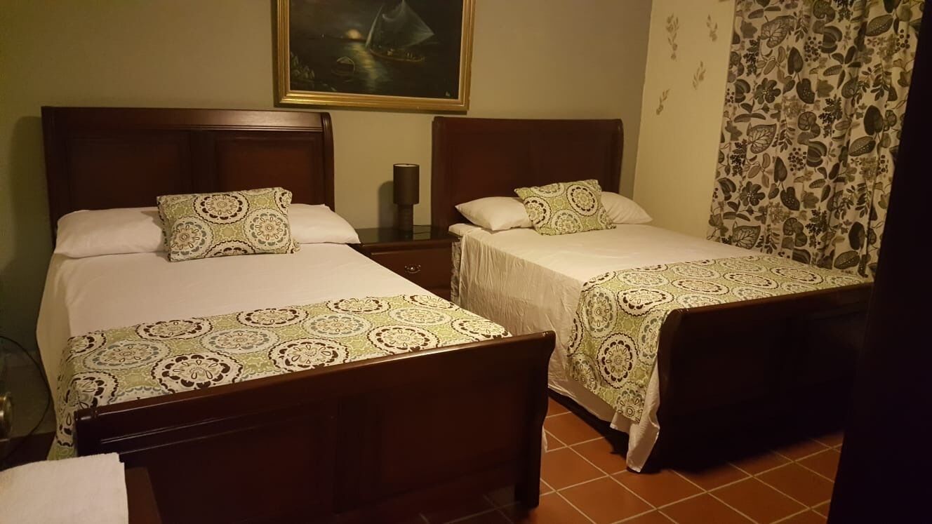 Een hotelkamer met twee eenpersoonsbedden, elk voorzien van een beddekleed met dessin en passende kussens. Daartussen staat een nachtkastje met een lamp, en aan de linker muur boven het hoofdeinde hangt een schilderij.