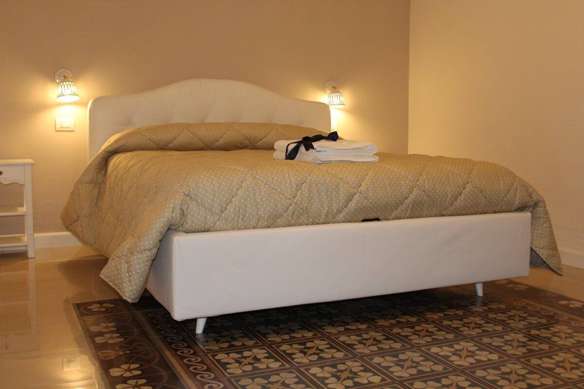 A neatly made bed with a quilted cover, a folded towel, and a headboard. There are two wall-mounted light fixtures on either side of the bed.