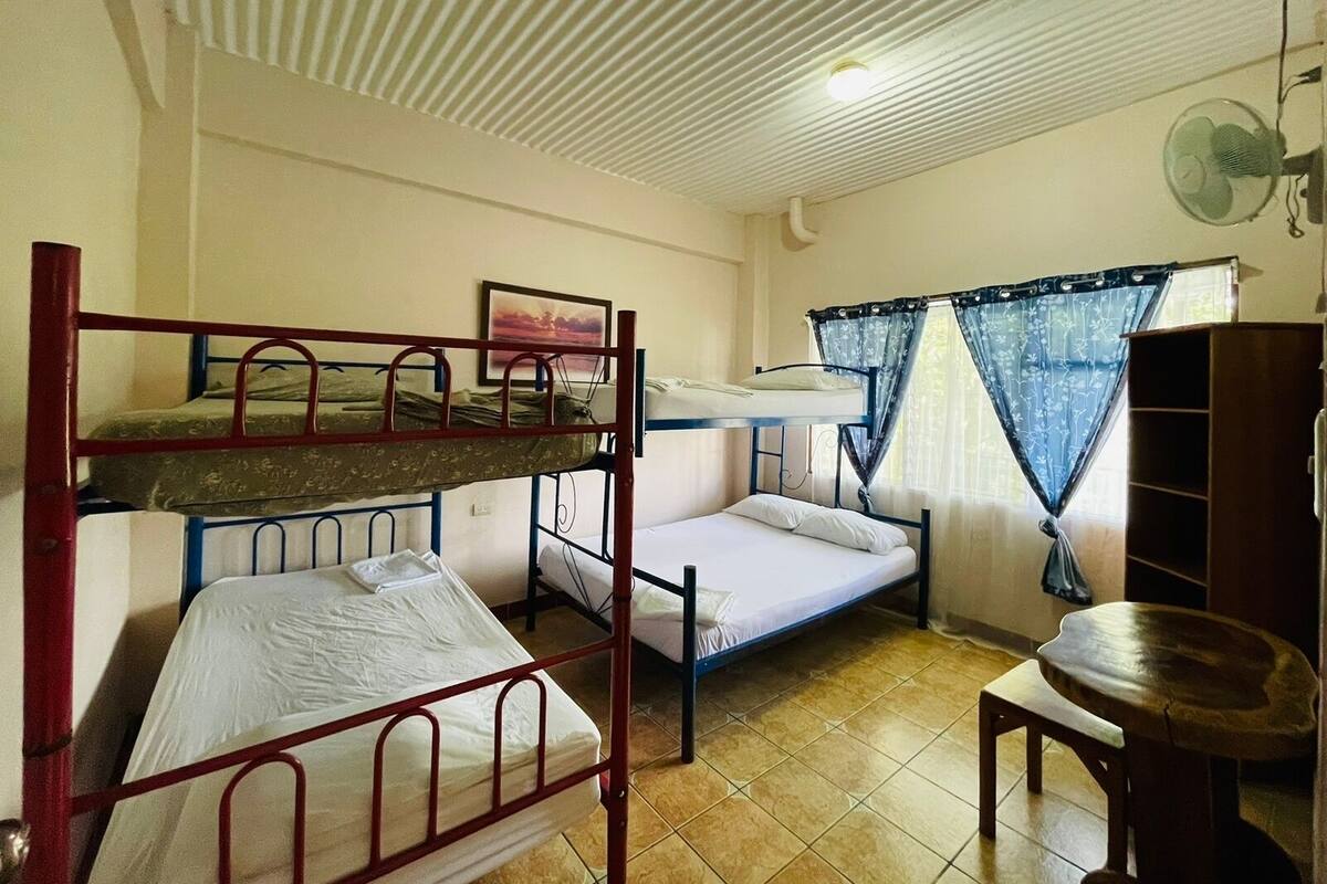 Un dormitorio con literas, un ventilador, una ventana con cortinas y una mesita.