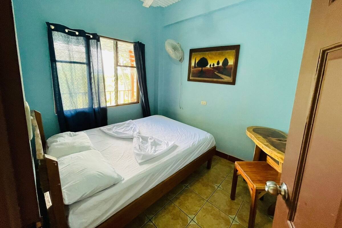 Una habitación con cama, escritorio, ventana con cortinas, ventilador y un cuadro en la pared.
