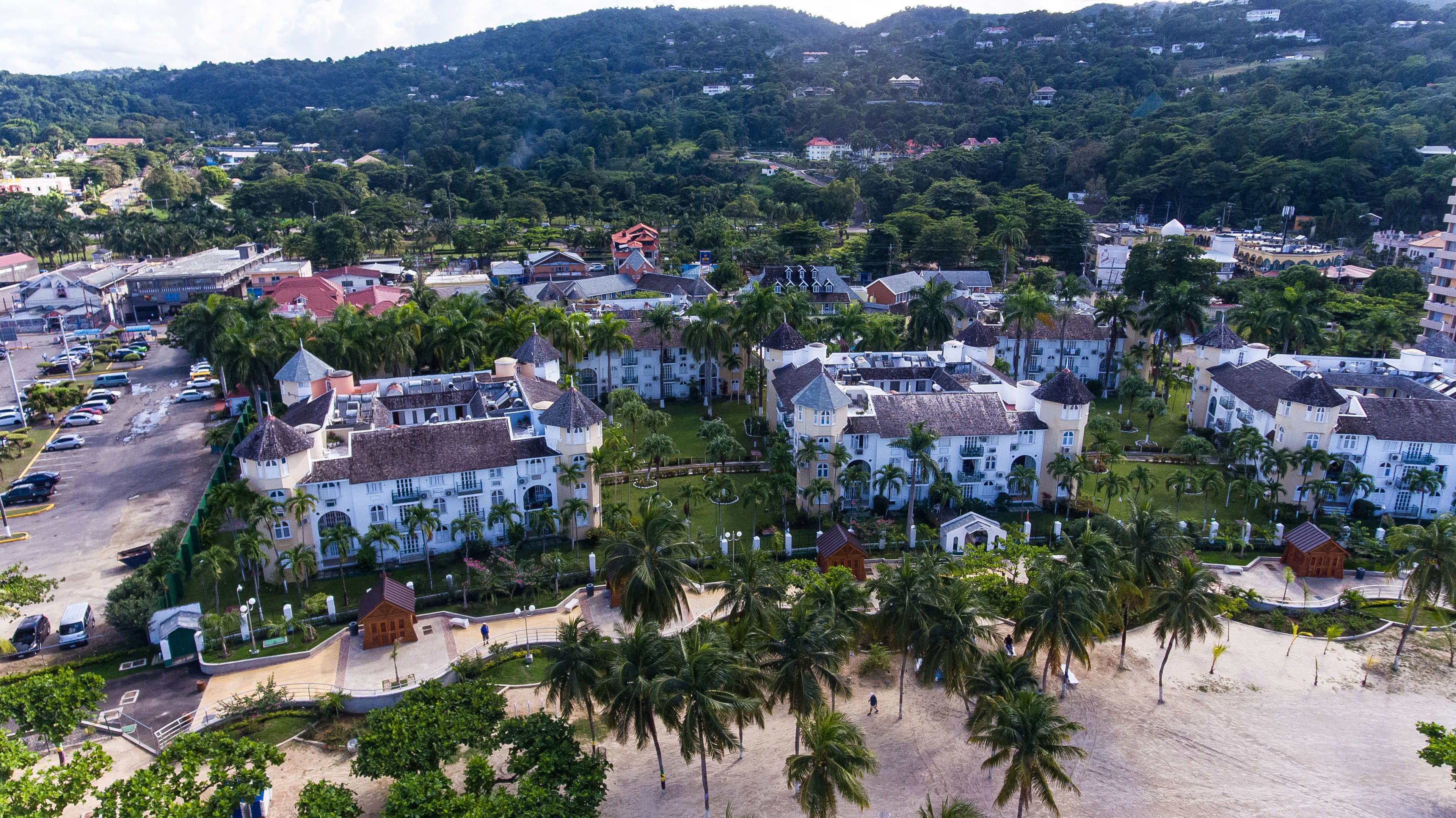 Vista aérea de una zona turística costera con varios edificios, incluyendo una estructura blanca destacada con techo rojo, rodeada de palmeras y una playa de arena.