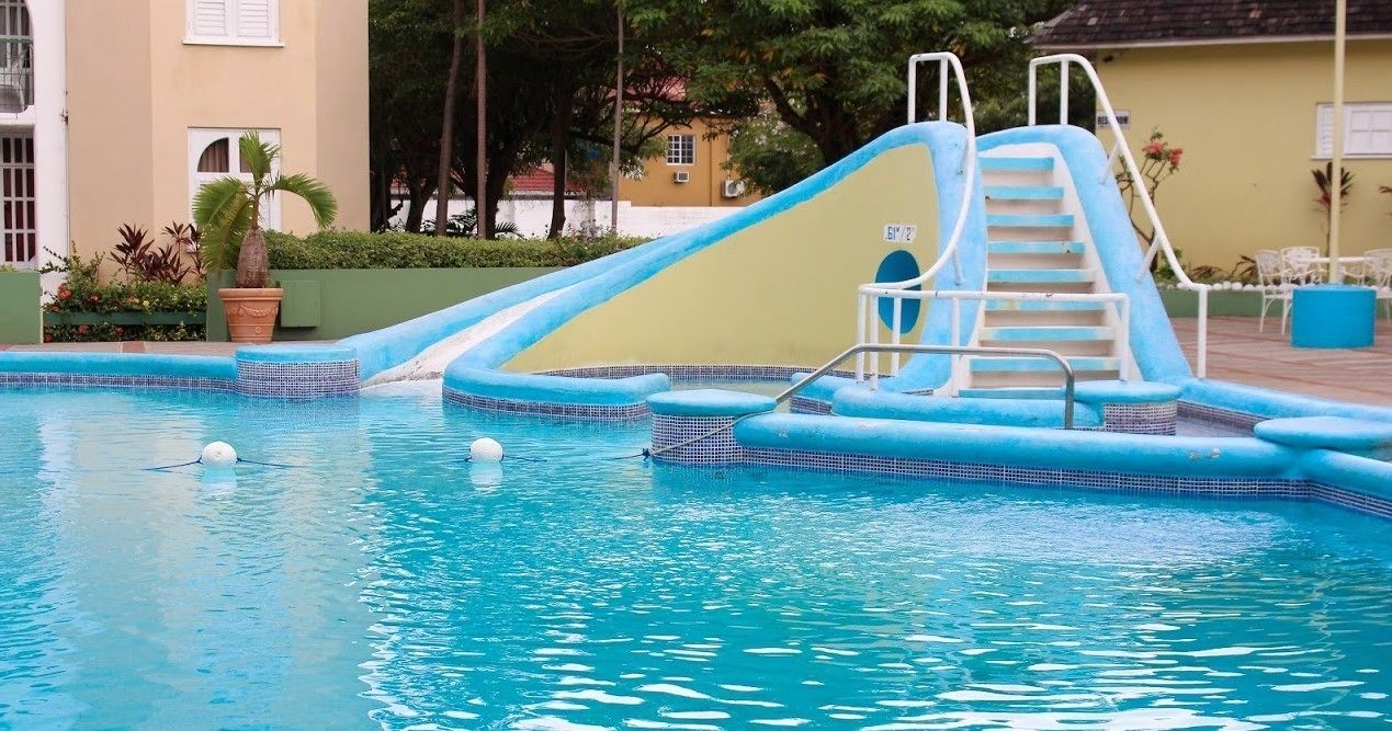 Una piscina con un tobogán azul y escaleras, rodeada de un suelo de baldosas y vegetación.