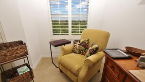 Ein Zimmer mit einem gelben Sessel, einem kleinen Holznachttisch, einem Korbnachttisch, einer Holzkommode und einem Fenster mit Jalousien.