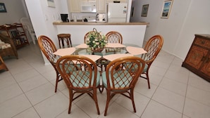 Un comedor con una mesa de vidrio rodeada de sillas de mimbre, una cocina al fondo y piso de baldosas.