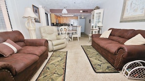 Ein Wohnzimmer mit zwei braunen Ledersofas, einem weißen Sessel und einem Esstisch mit Stühlen. Im Hintergrund ist eine Küche zu sehen.