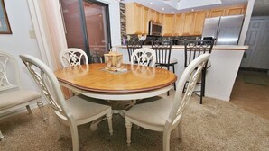 Ein Essbereich mit einem runden Holztisch und weißen Stühlen, eine Küche mit Holzschränken und Edelstahlgeräten sowie ein Teppichboden.