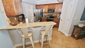 A kitchen with wooden cabinets, a stainless steel refrigerator, a microwave, and a granite countertop. There are two white bar stools and a small wooden table with a basket. - Oceanwalk 18-406 Apartment 2 (New Smyrna Beach)