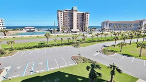 A parking lot with marked spaces, palm trees, and a view of a hotel and ocean. - Oceanwalk 18-406 Apartment 2 (New Smyrna Beach)