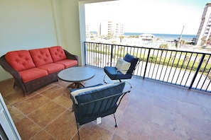 A balcony with a red sofa, a round table, and two chairs. The balcony overlooks a coastal area with buildings and a view of the sea.