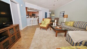 A living room with a sofa, chairs, a coffee table, a TV, and a kitchen area in the background. - Oceanwalk 18-406 Apartment 2 (New Smyrna Beach)
