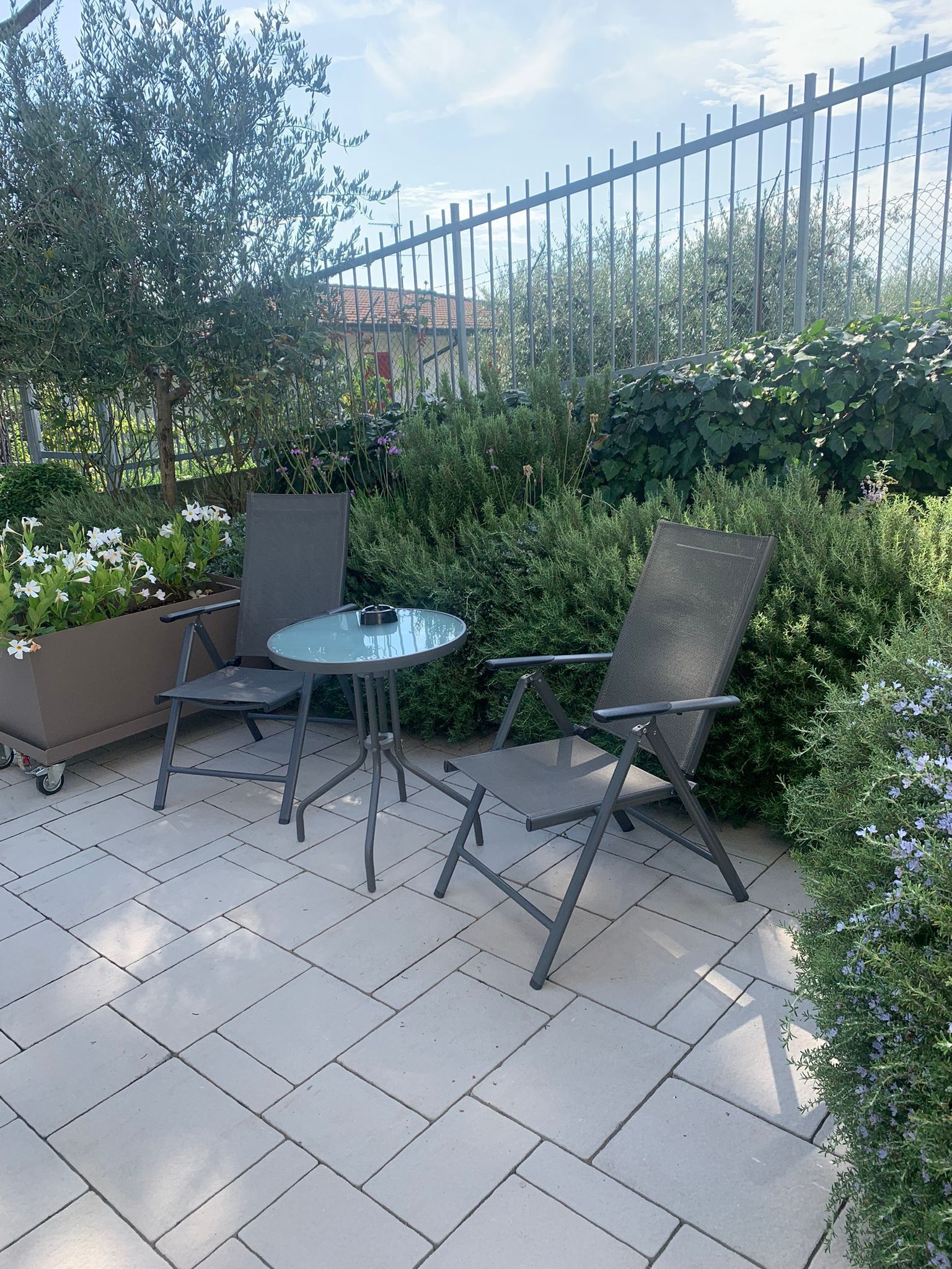 An outdoor patio area with a round glass table and two metal chairs, surrounded by potted plants and a metal fence.