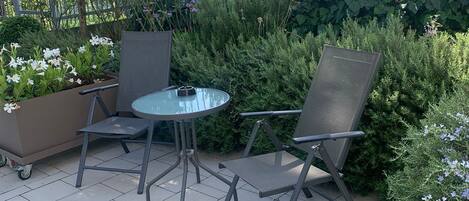 An outdoor patio area with a round glass table and two metal chairs, surrounded by potted plants and a metal fence.