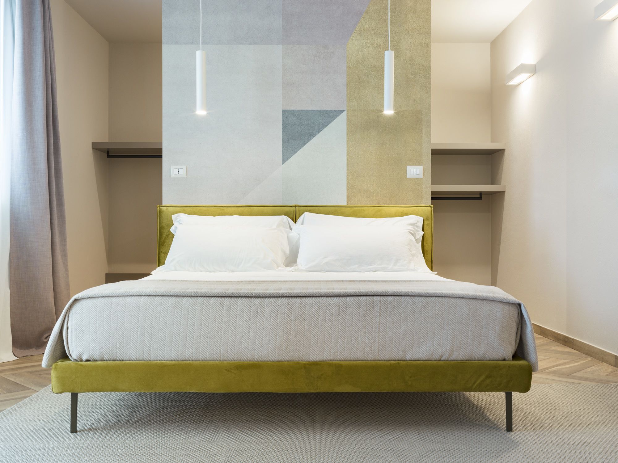 Une chambre moderne avec un lit recouvert d’un tissu vert, du linge de lit blanc et des luminaires suspendus.