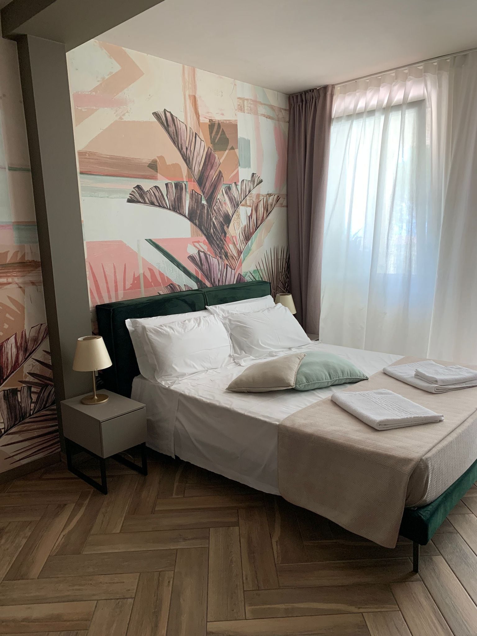 A hotel room with a bed, a nightstand, a lamp, a window with curtains, and a wall with a tropical leaf mural.