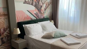 A hotel room with a bed, a nightstand, a lamp, a window with curtains, and a wall with a tropical leaf mural.