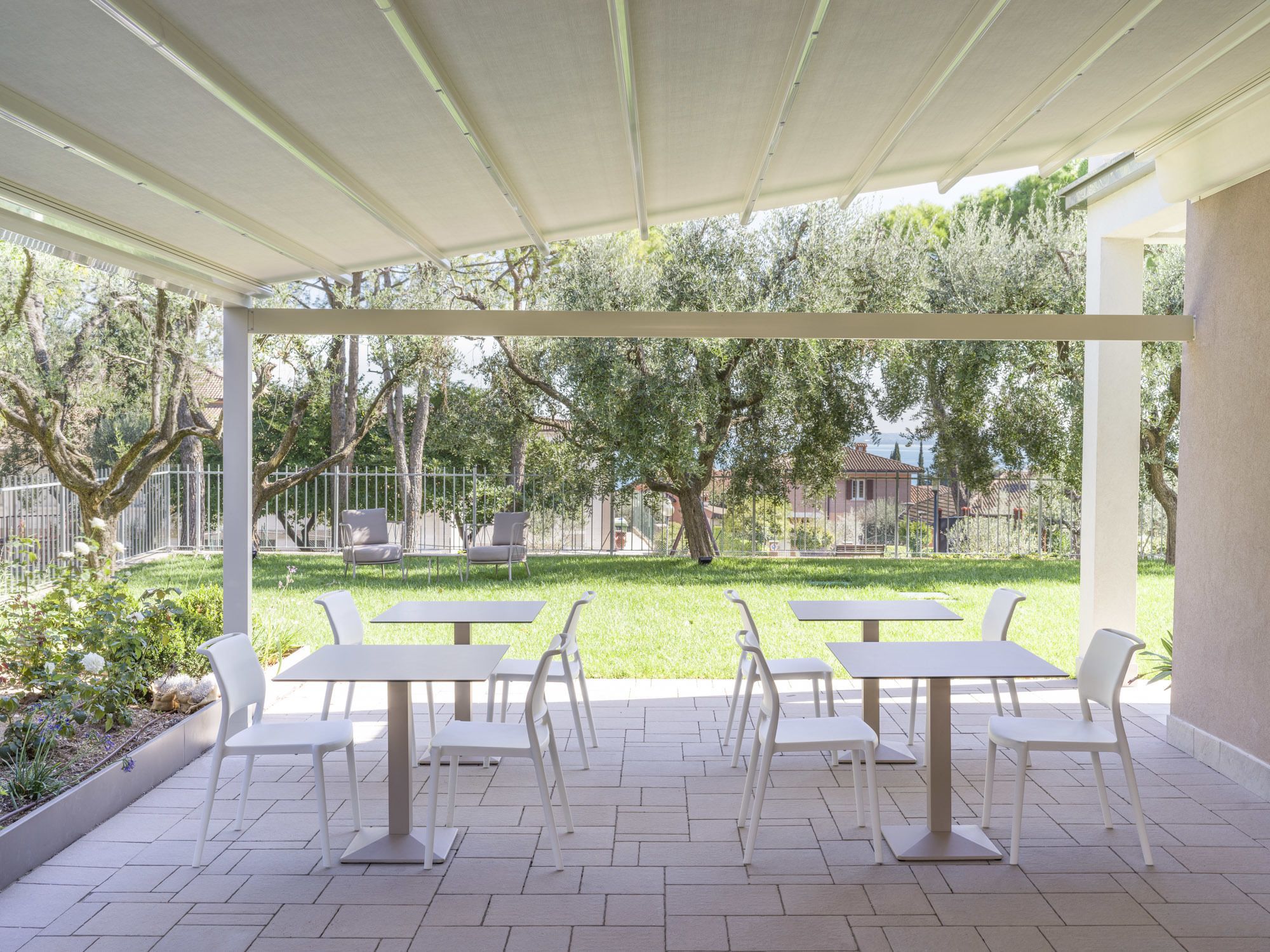 An outdoor patio with white tables and chairs, a pergola overhead, and a view of a grassy area with trees and a fence.