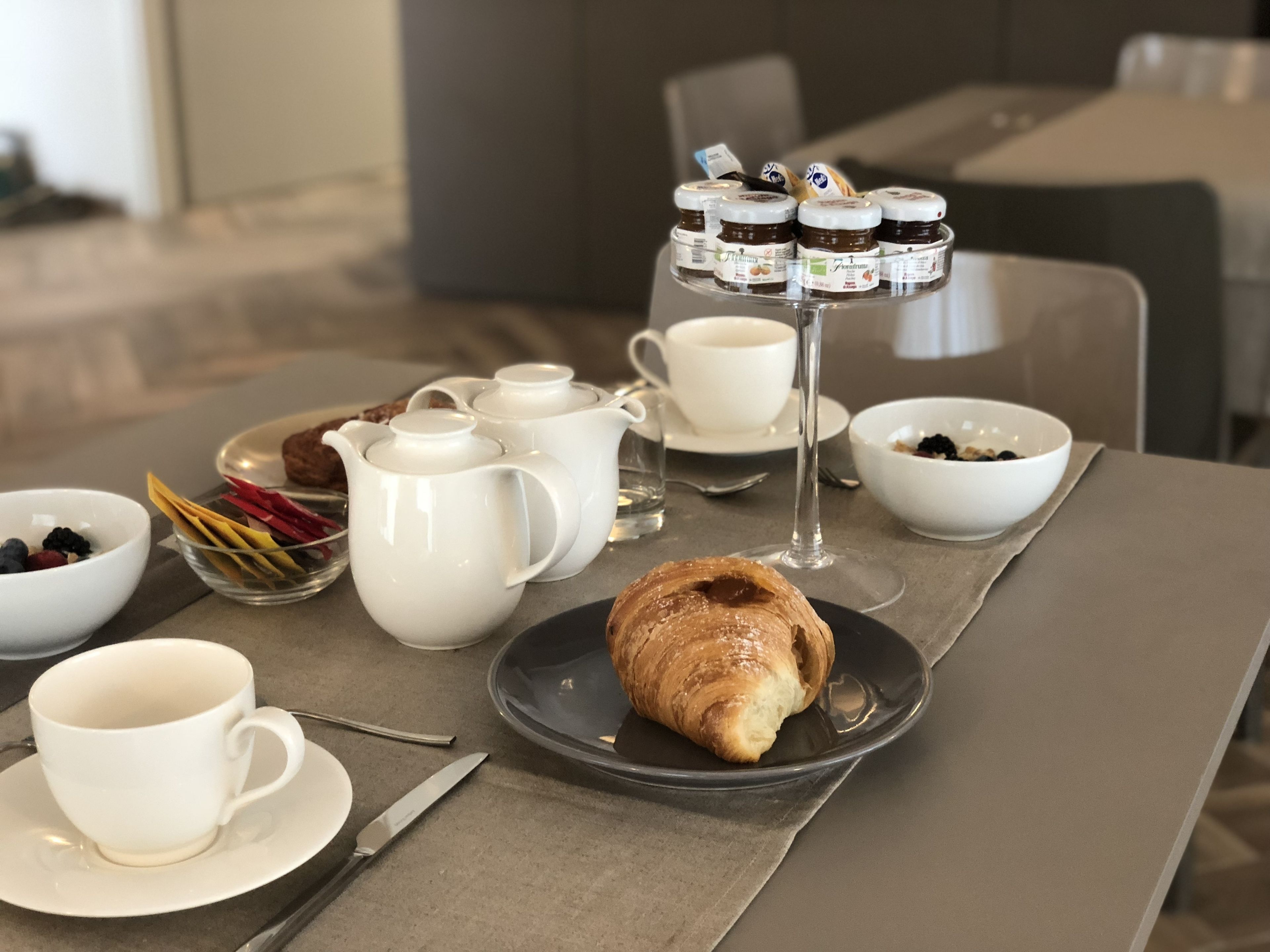A breakfast setting with croissants, coffee cups, and jam jars on a table.