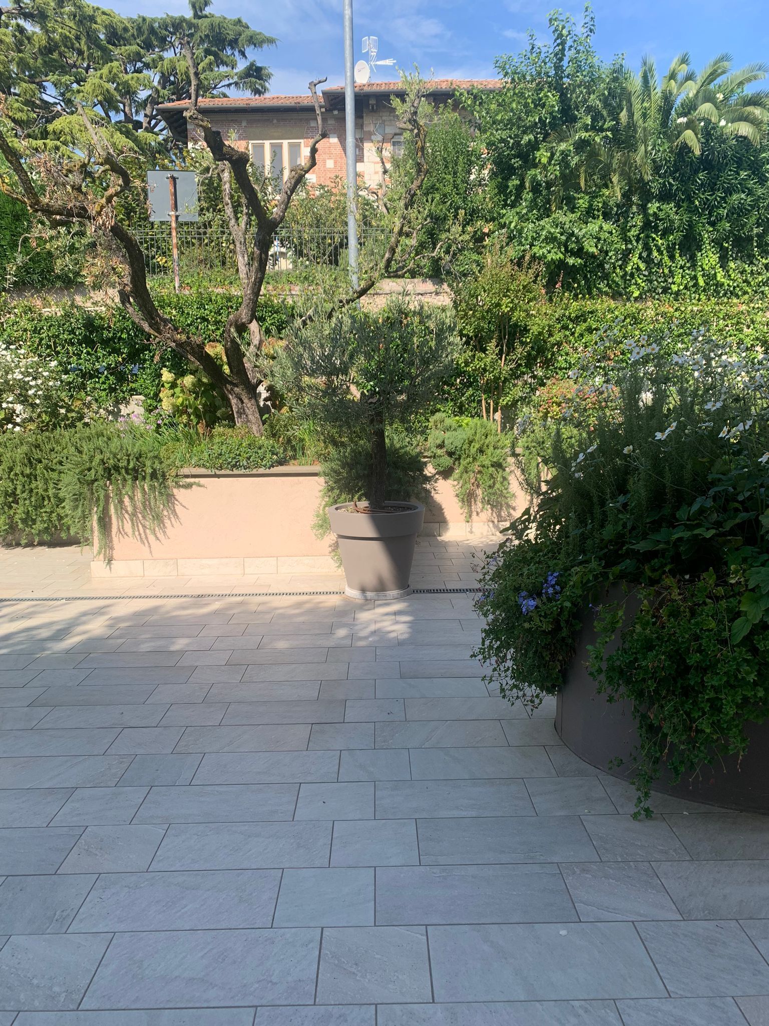 En anlagt sti med et metalhegn, omgivet af frodig vegetation og en bygning med et tegltag i baggrunden.