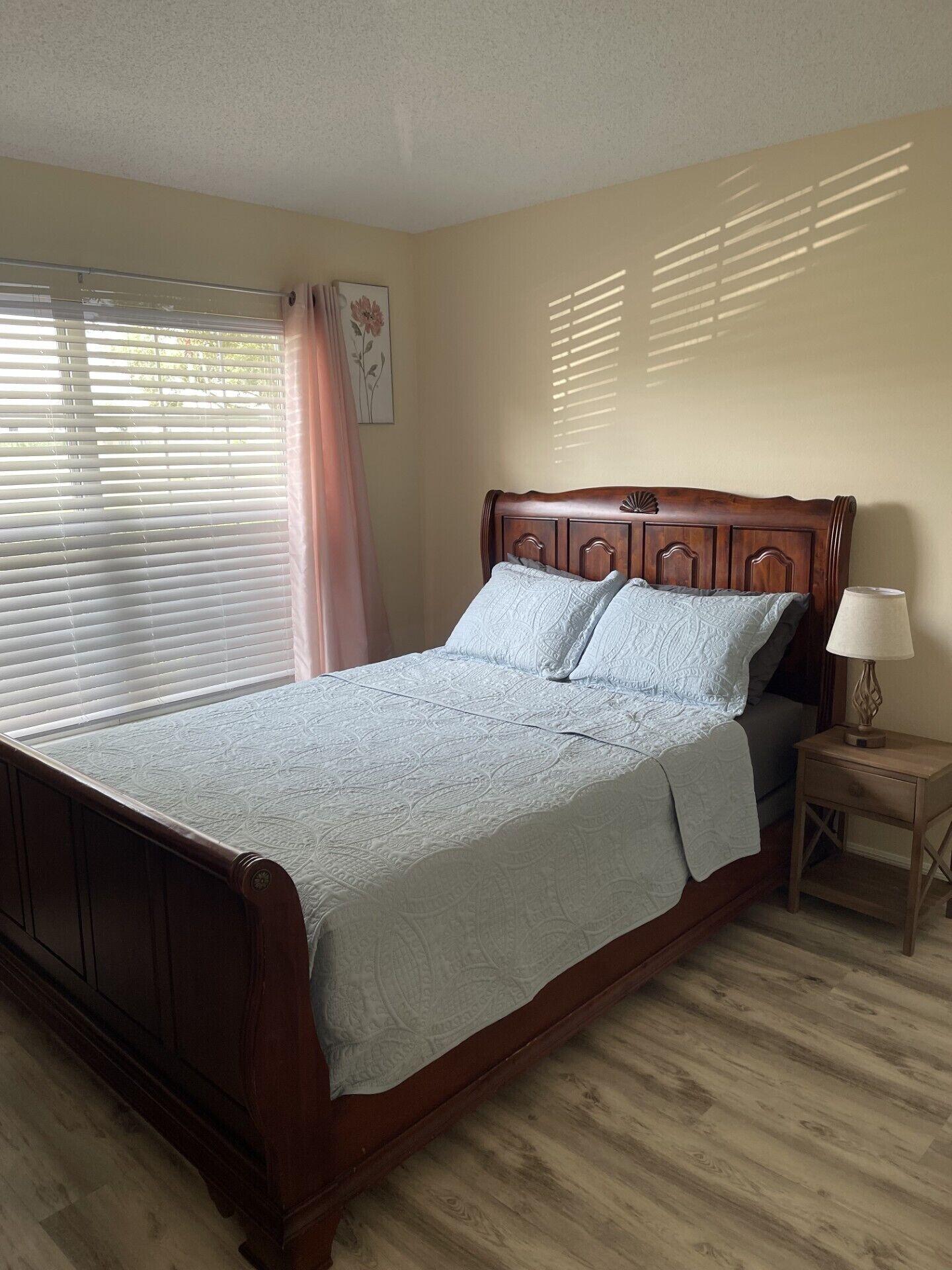 A bedroom with a wooden bed, a bedside table with a lamp, and a window with blinds.
