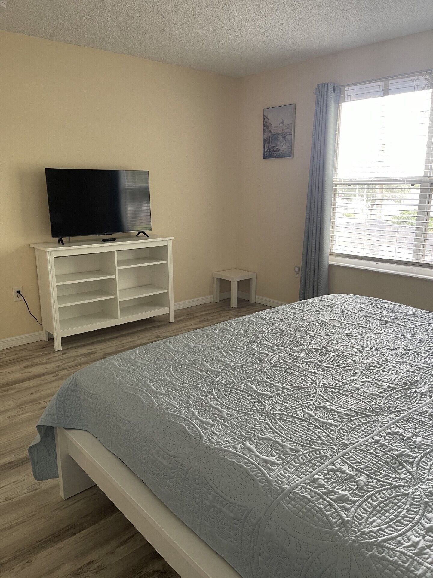 A bedroom with a bed, a TV on a white stand, a small table, and a window with blinds.