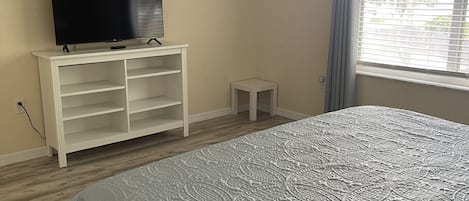 A bedroom with a bed, a TV on a white stand, a small table, and a window with blinds.