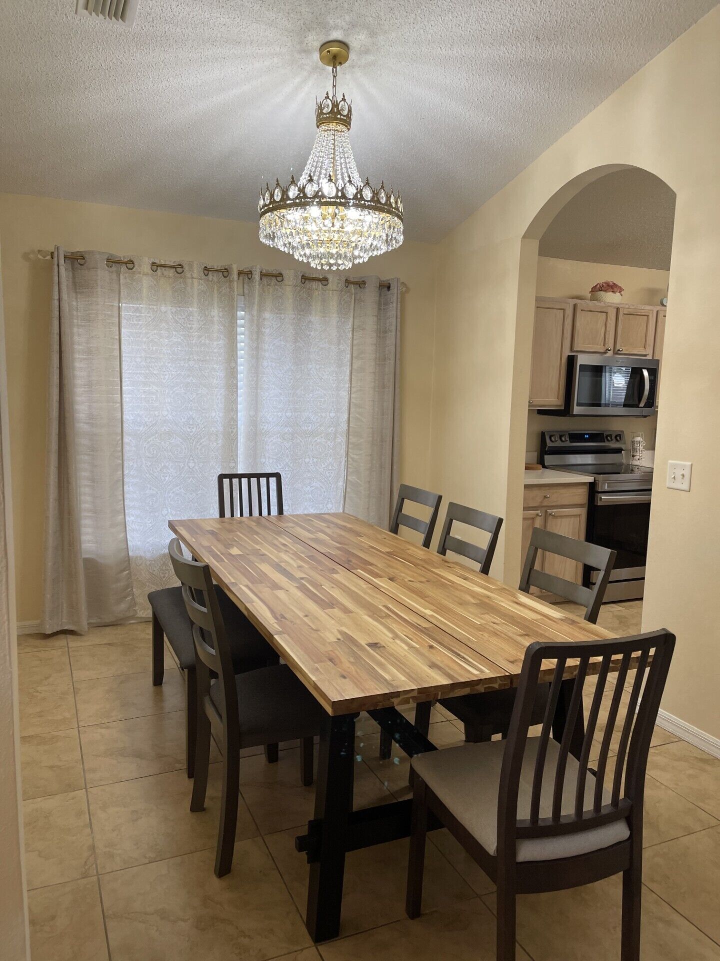 A dining room with a wooden table and chairs, a chandelier, and a kitchen in the background.