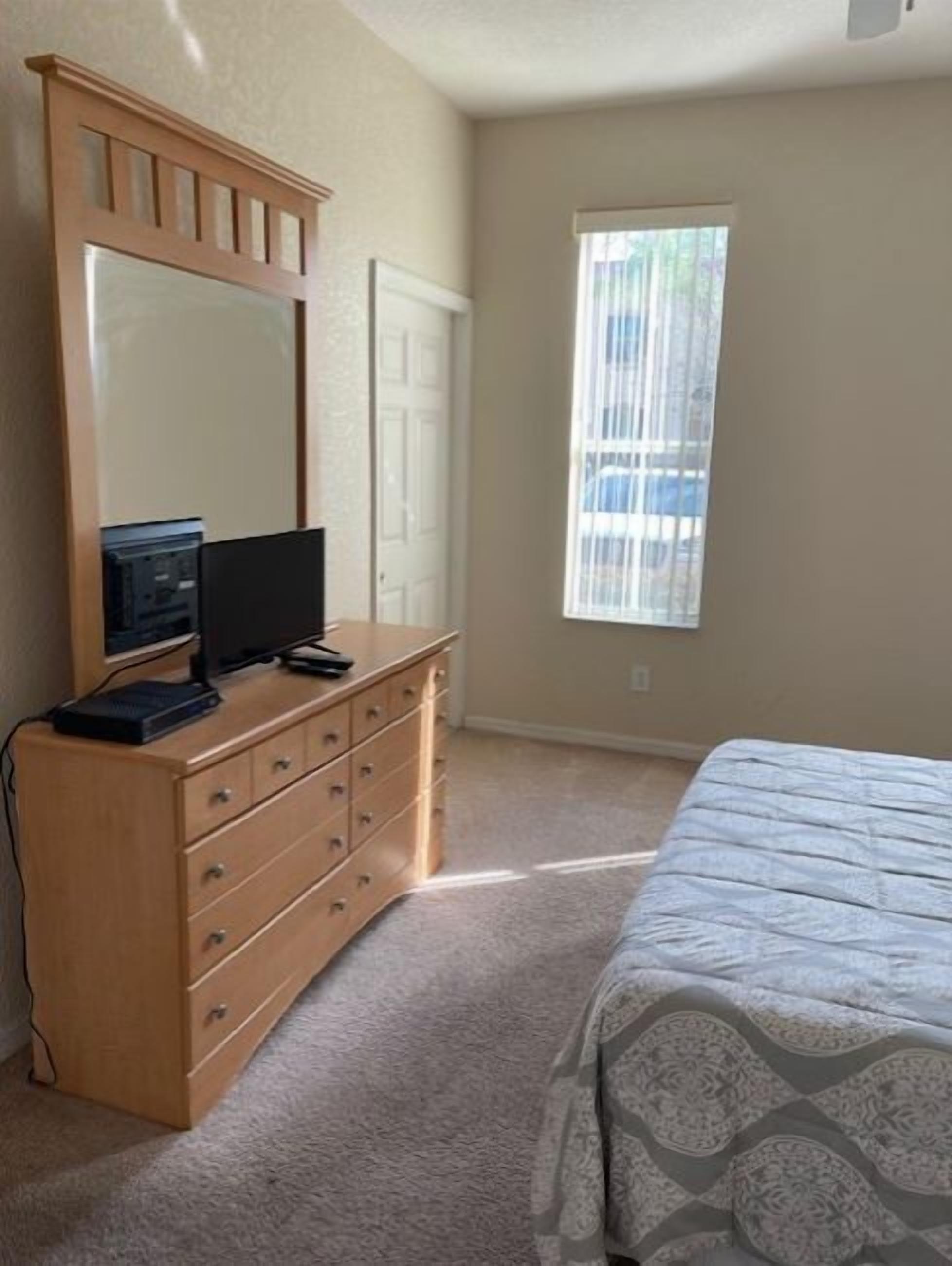 Un dormitorio con un aparador de madera, un televisor, una cama con ropa de cama estampada, una ventana con persianas y una puerta.
