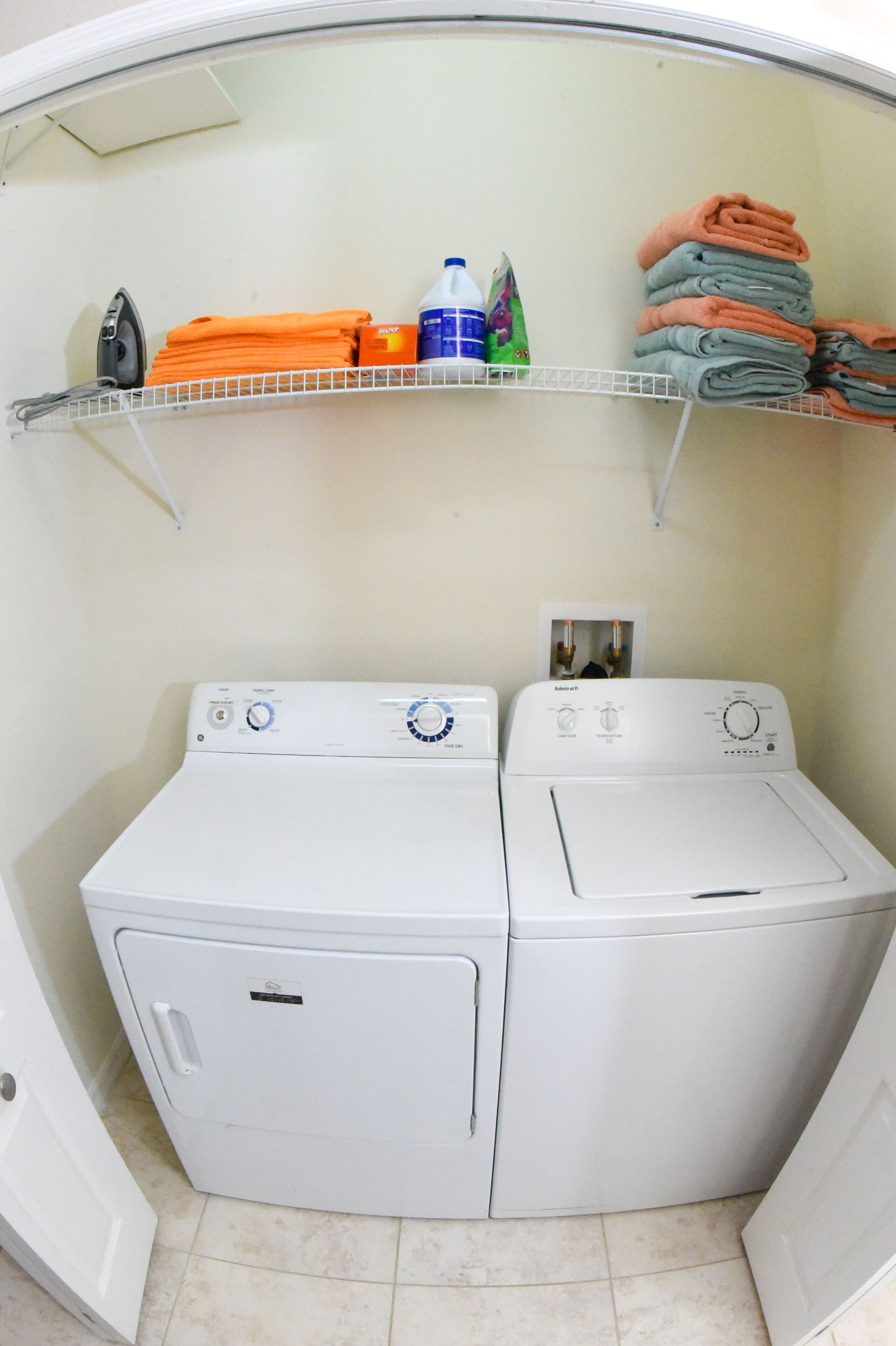 A laundry room with two white washing machines, a shelf with folded towels and laundry supplies, and a door to the left.