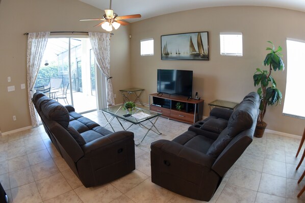 A living room with two dark brown sofas, a glass coffee table, a TV on a wooden stand, a ceiling fan, and a potted plant.