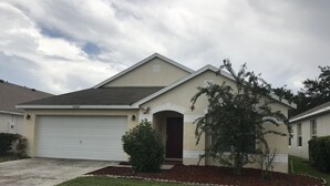 A single-story house with a garage, a front door, and a small front yard with a tree and shrubs.