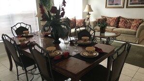 A dining area with a wooden table set for four, surrounded by chairs. There are two potted plants, a lamp, and framed artwork on the walls.