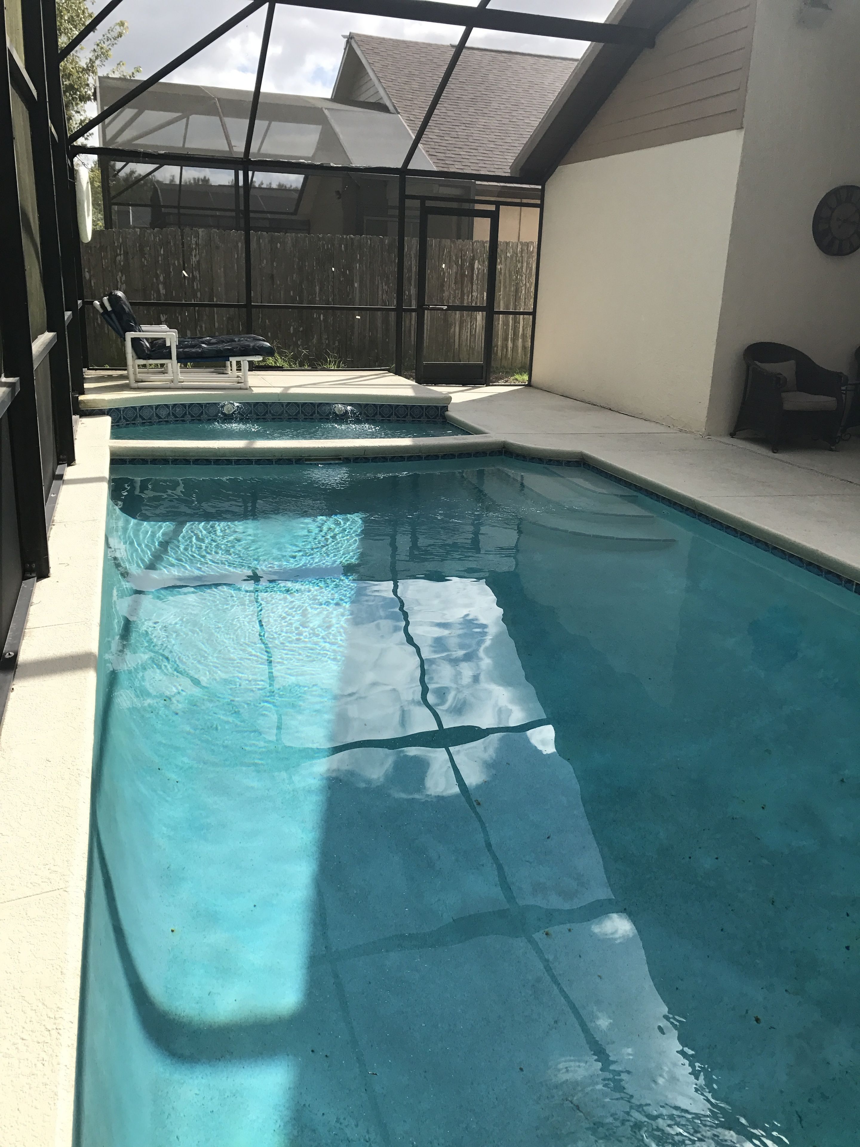 A clear swimming pool with a tiled edge, a wooden fence in the background, and a covered patio area with chairs.