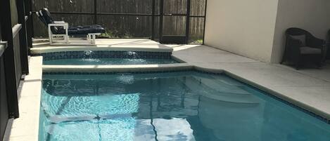 A clear swimming pool with a tiled edge, a wooden fence in the background, and a covered patio area with chairs.