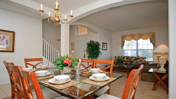 A dining area with a glass table, wooden chairs, and a chandelier. There is a staircase in the background leading to another room.