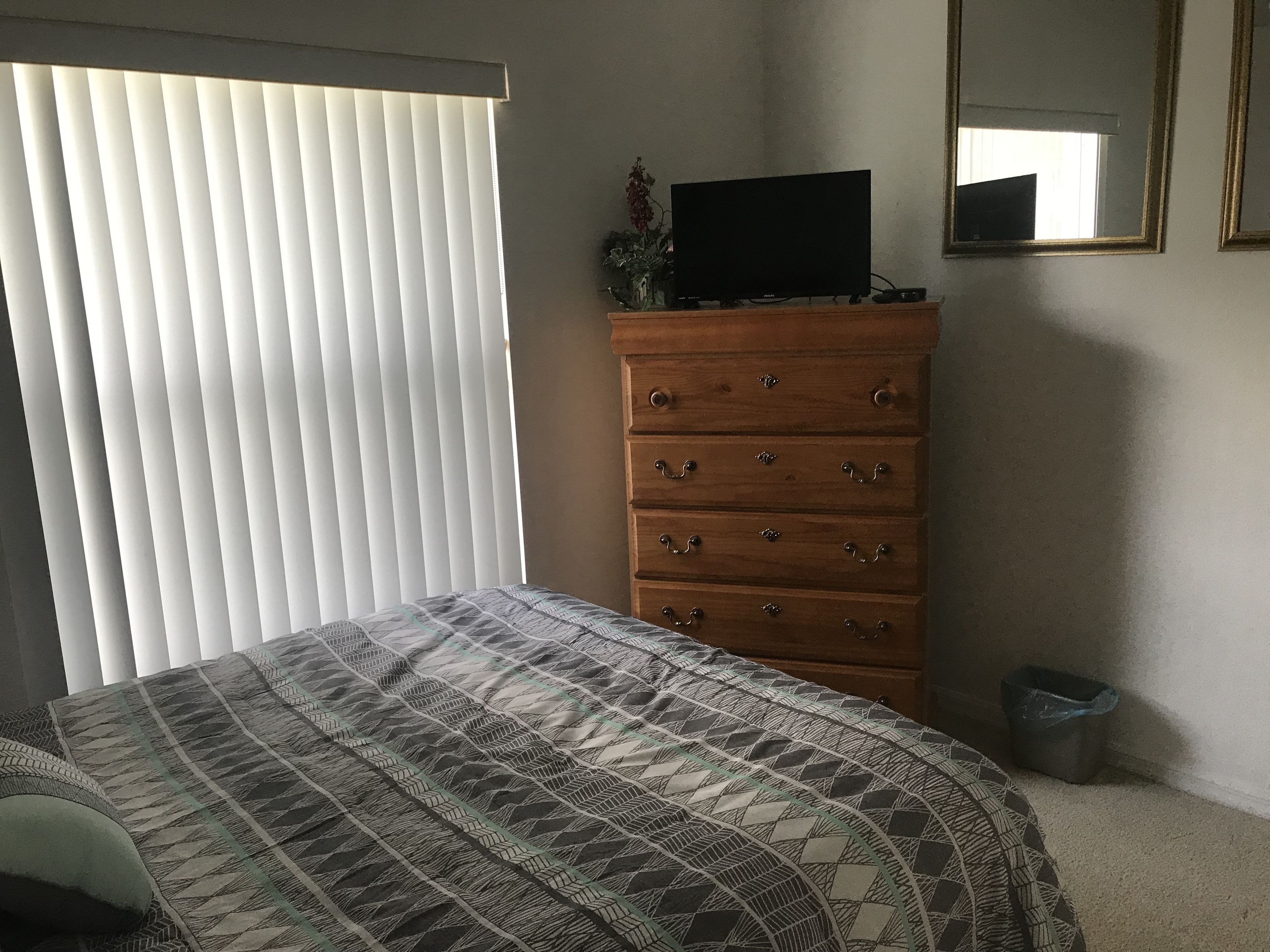 Une chambre à coucher comprenant un lit, une commode, un téléviseur et une fenêtre avec des stores.