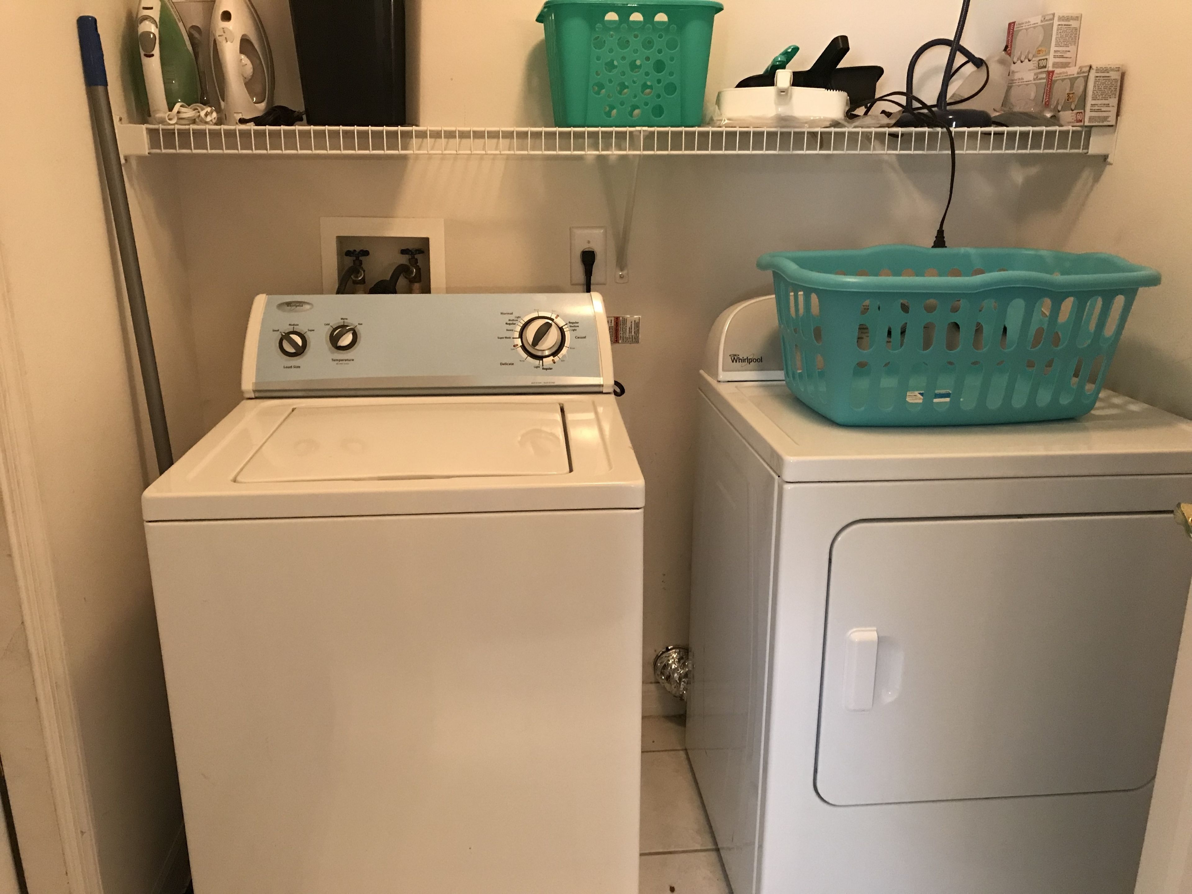Un lavadero pequeño con lavadora y secadora de carga frontal, un estante sobre la secadora con productos de limpieza, y una cesta de ropa del color turquesa sobre la secadora.