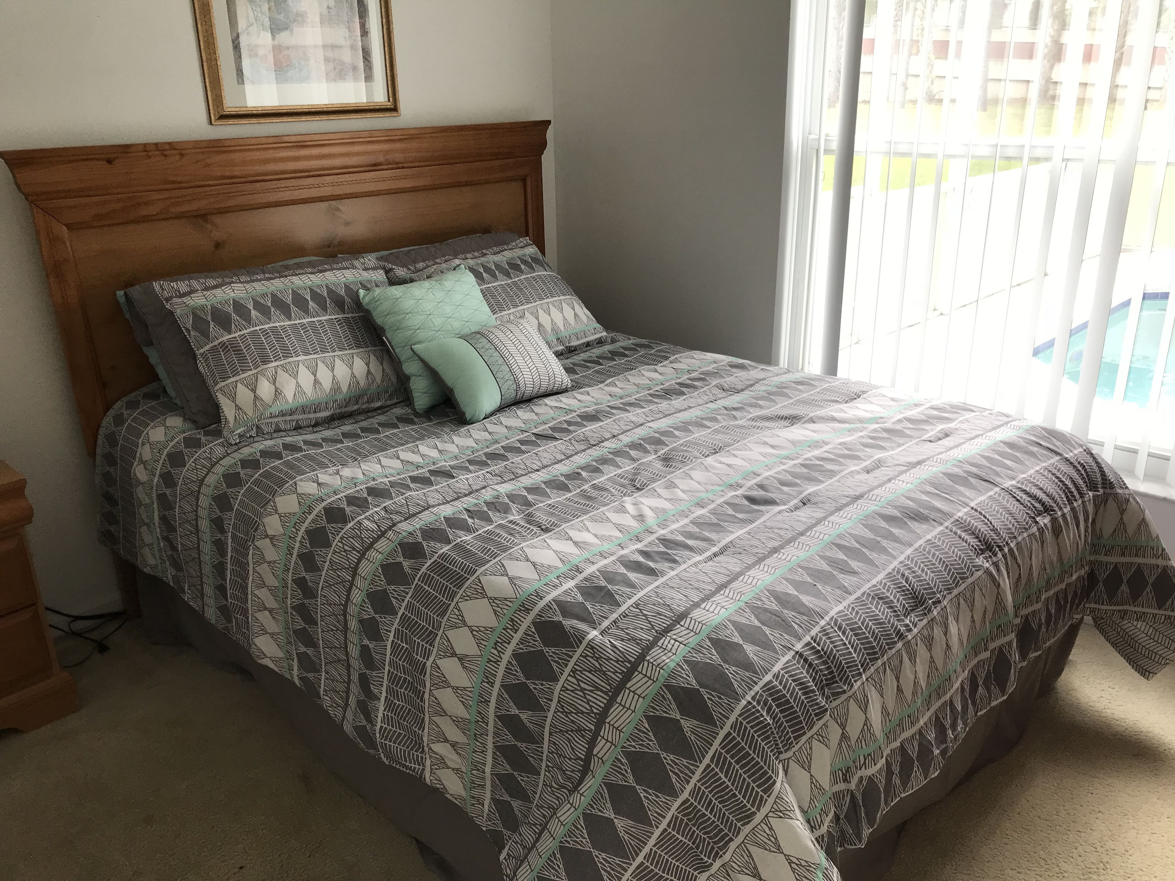 A bedroom with a wooden headboard, a patterned bedspread, and a window with blinds.