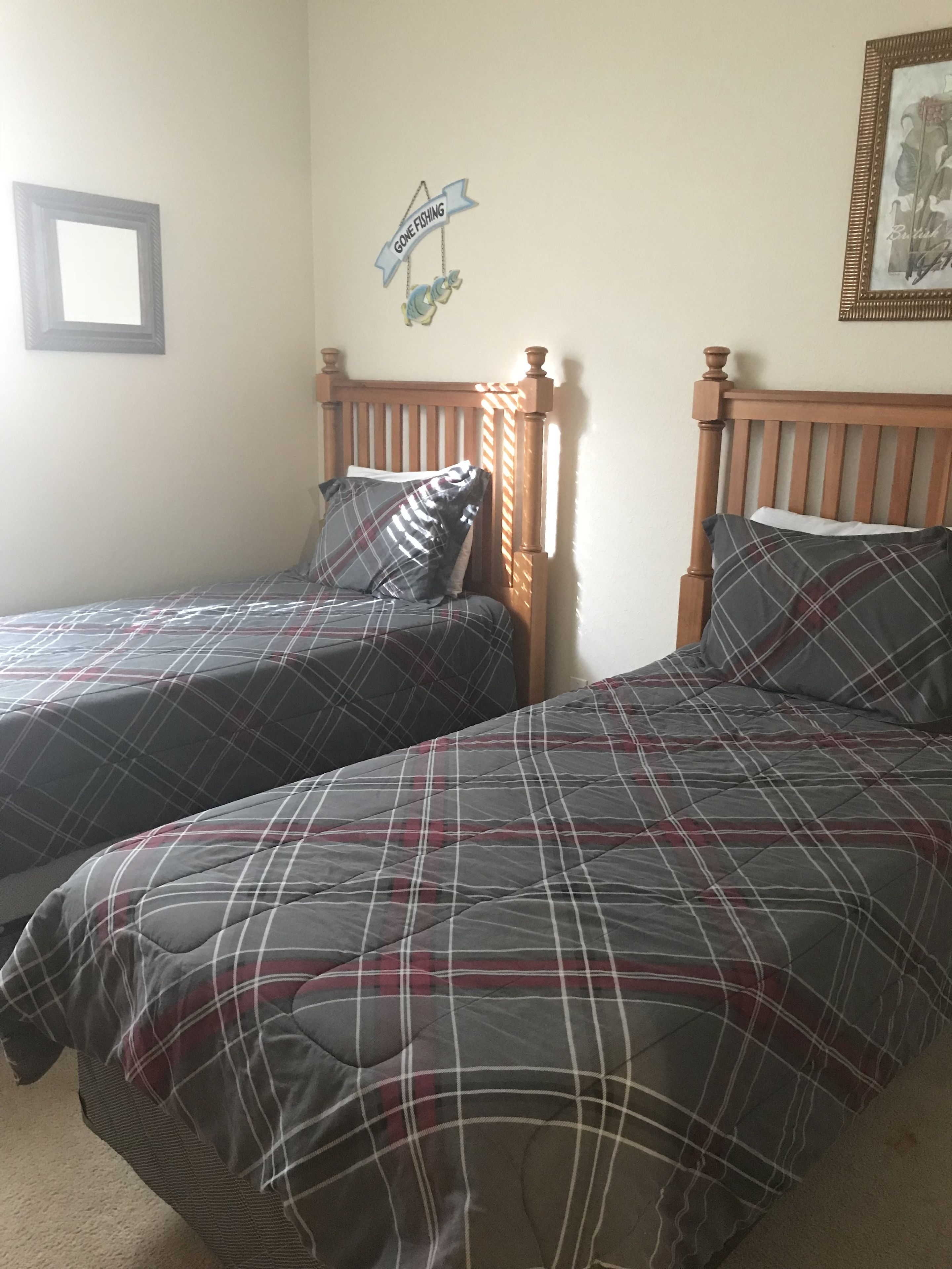 Une chambre avec deux lits, chacun recouvert d’une couverture à carreaux et agrémenté d’oreillers. Un tableau encadré est accroché au mur, et une pancarte indique : « En train de pêcher ».
