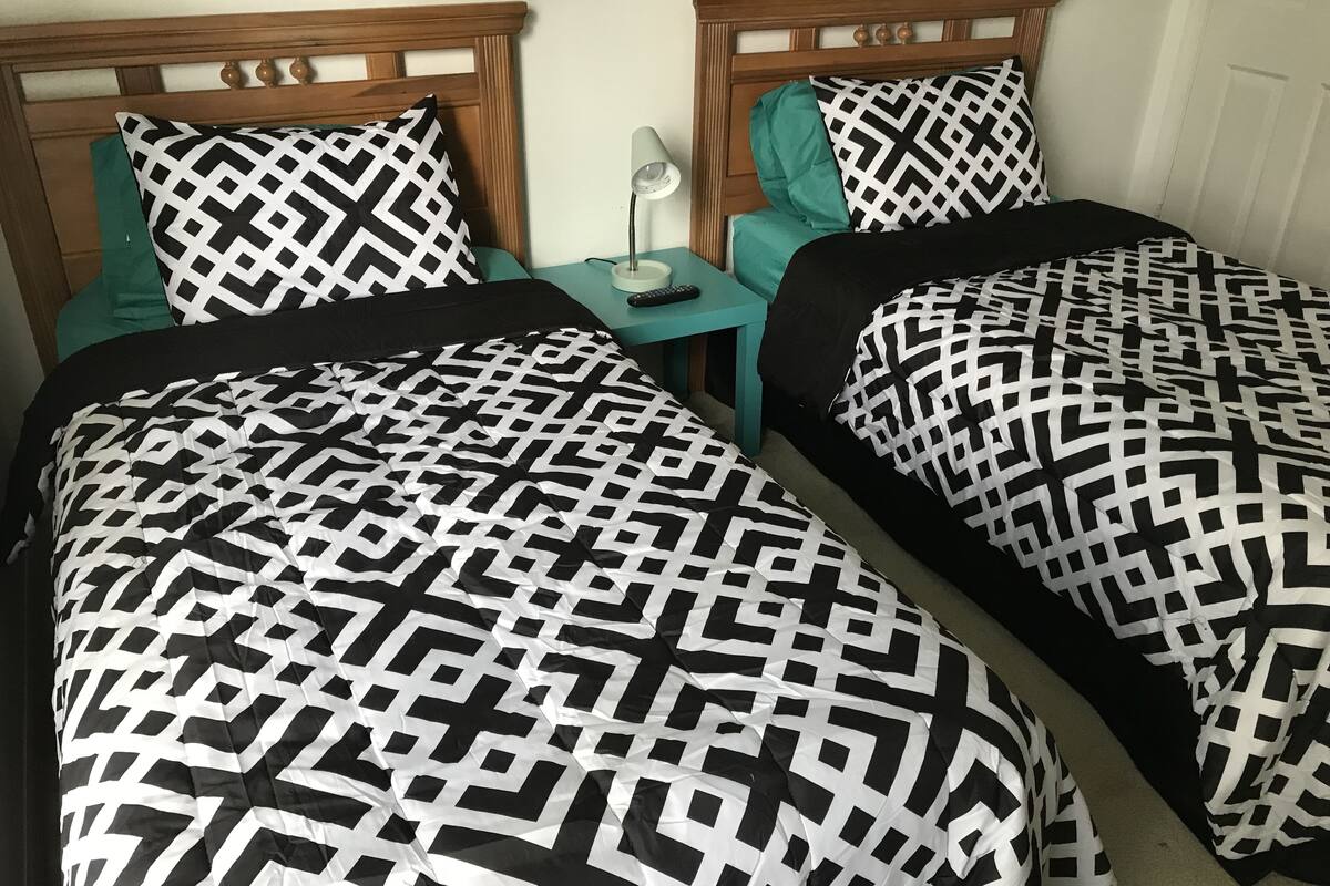 Una habitación con dos camas, cada una con cabecero de madera y ropa de cama de estampado blanco y negro. Hay una mesita de noche color verde azulado con una lámpara y un control remoto.