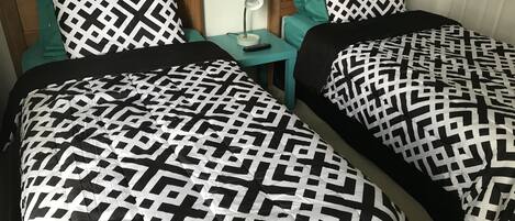 Una habitación con dos camas, cada una con cabecero de madera y ropa de cama de estampado blanco y negro. Hay una mesita de noche color verde azulado con una lámpara y un control remoto.
