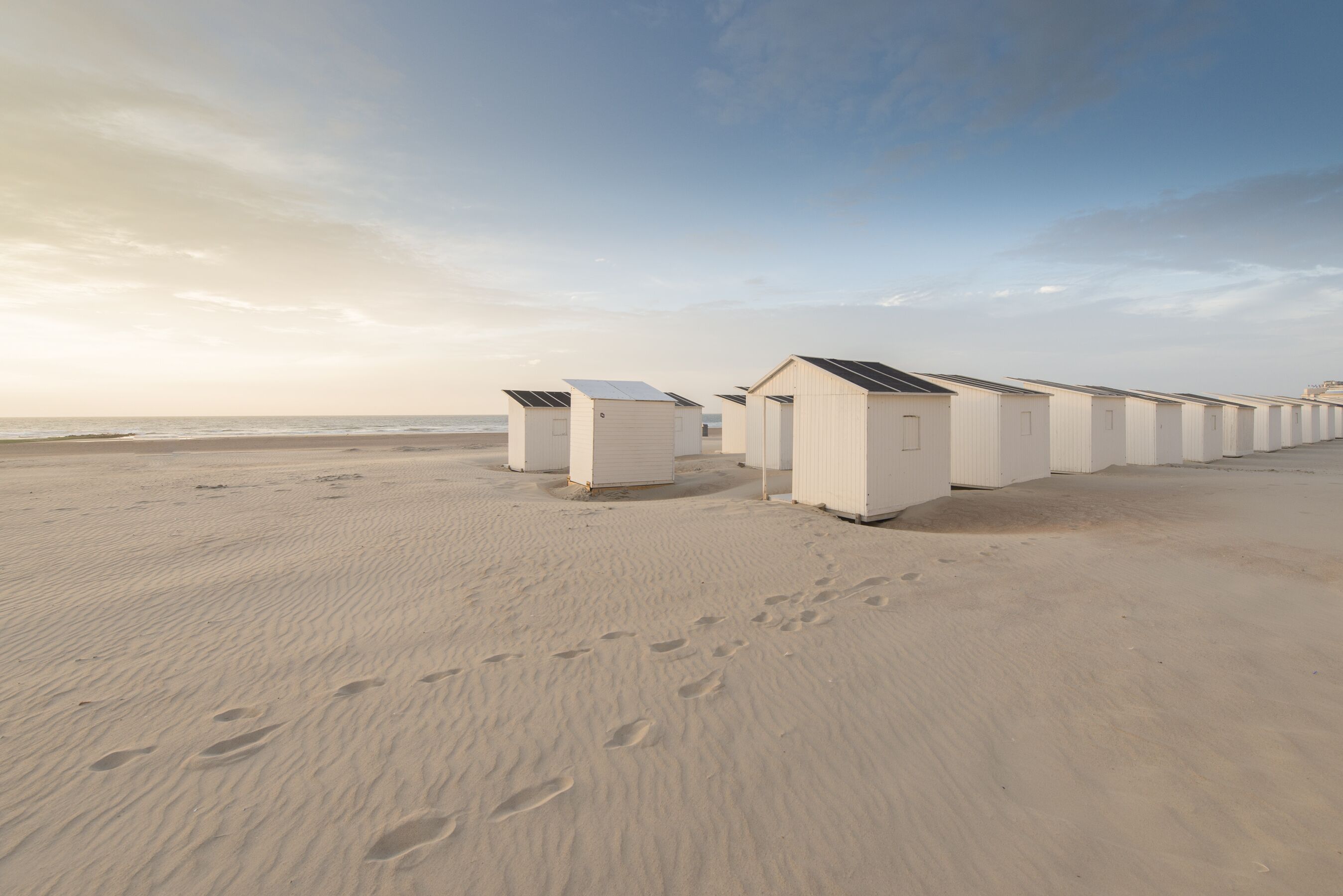 Een rij witte strandhutten met zonnepanelen op de daken, langs een zandstrand met voetafdrukken die ernaartoe leiden.